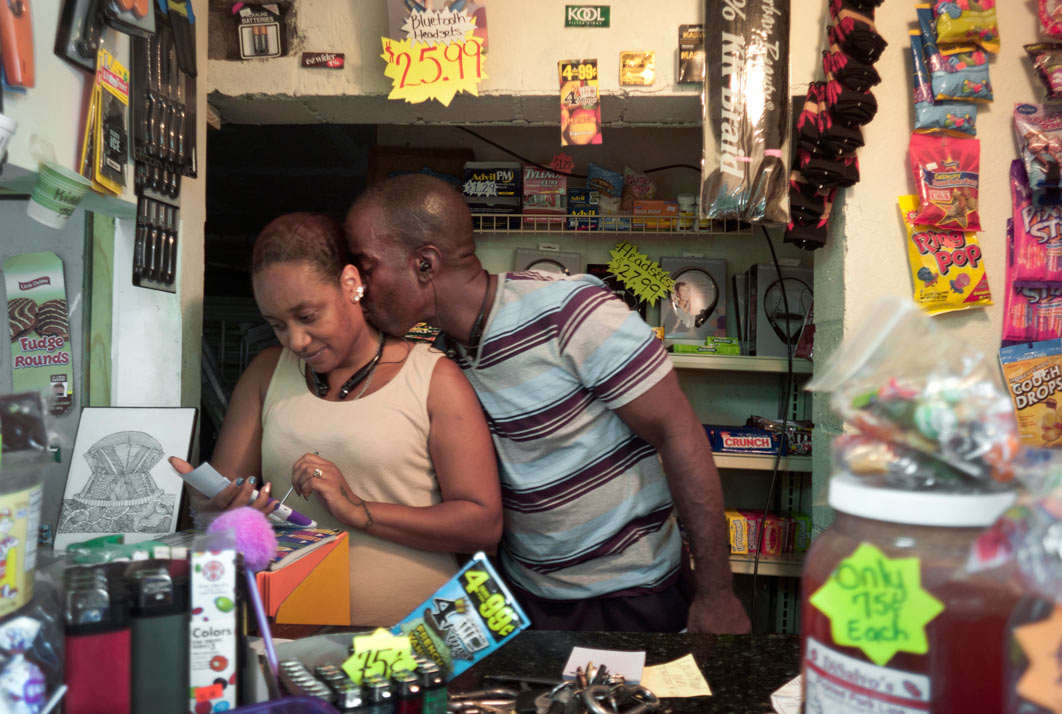Planting a quick kiss on his wife's neck, Burnell Cotlon and his wife Keasha work behind the counter of their Lower Ninth Ward Market. Using their savings, they bought the flood building that was destroyed by Hurricane Katrina. Burnell and Keasha refer to the store as a mini-Costco because it has a little bit of everything. His mother, Lillie Cotlon, works in the concession stand and helps stock shelves. Before the store opened, Keasha would drive elderly residents or unemployed mothers with children miles to the nearest grocery store who otherwise would have had to ride long distances on slow city buses just to buy food. 
